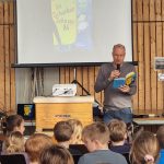Ein Mann steht in einer Turnhalle vor einer Leinwand und liest aus einem blauen Buch vor. Er hält ein Mikrofon in der Hand und trägt ein graues Langarmshirt und eine Jeans. Auf der Projektionsleinwand hinter ihm ist das Cover des Kinderbuches „Die Schurken-Schnapp-AG" von Martin Muser zu sehen, das als „Buch des Monats" ausgezeichnet ist. Vor ihm sitzen zahlreiche Grundschulkinder auf Stühlen und hören aufmerksam zu. Im Vordergrund ist ein Turnkasten der Marke Sport-Thieme zu sehen, auf dem ein Beamer steht.