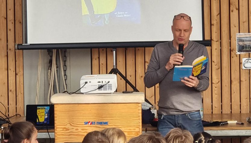 Ein Mann steht in einer Turnhalle vor einer Leinwand und liest aus einem blauen Buch vor. Er hält ein Mikrofon in der Hand und trägt ein graues Langarmshirt und eine Jeans. Auf der Projektionsleinwand hinter ihm ist das Cover des Kinderbuches „Die Schurken-Schnapp-AG" von Martin Muser zu sehen, das als „Buch des Monats" ausgezeichnet ist. Vor ihm sitzen zahlreiche Grundschulkinder auf Stühlen und hören aufmerksam zu. Im Vordergrund ist ein Turnkasten der Marke Sport-Thieme zu sehen, auf dem ein Beamer steht.
