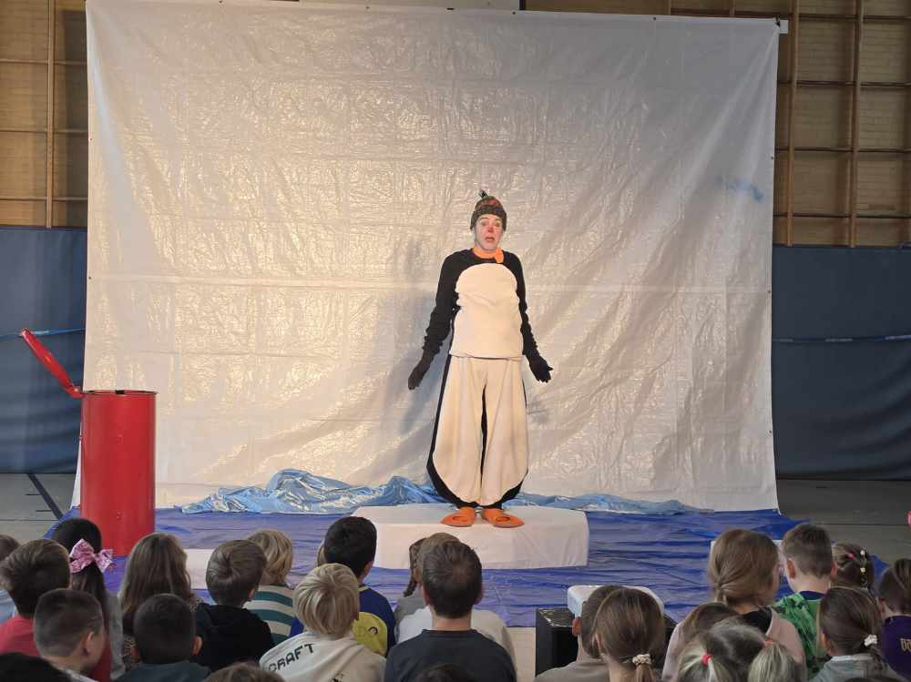 Ein Schauspieler im Pinguinkostüm steht auf einer Bühne in der Turnhalle, davor sitzen Kinder und schauen zu.