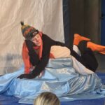 Der Pinguin aus dem Theaterstück liegt mit Mütze, Sonnenbrille und orangefarbenem Schal entspannt auf einem hellblauen Stoffhügel.